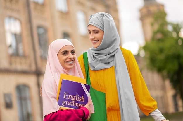 Petite amie avec un ami. Étudiant rayonnant portant un hijab rose tenant un livre anglais debout près d'un ami