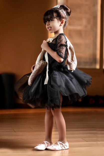 Une petite ballerine vêtue d'une robe de ballet noire et portant des chaussures de ballet sur son épaule