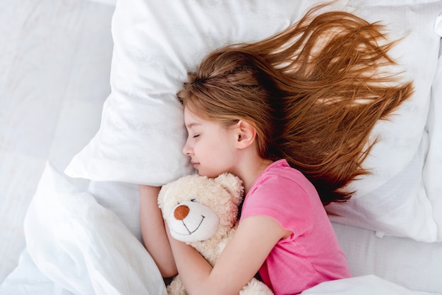 Petite belle fille allongée dans le lit étreignant un ours en peluche et dormant. Enfant préadolescent au repos portant un t-shirt rose et tenant un jouet. Portrait de jolie femme aux cheveux longs