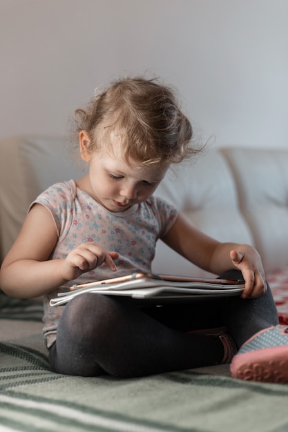 Petite belle fille assise avec une tablette sur le canapé et joue à des jeux. Isolement, maison, enfant