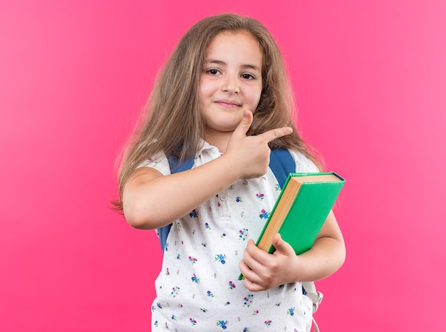 Petite belle fille aux cheveux longs avec sac à dos tenant un cahier regardant devant souriant pointant gaiement avec l'index sur le côté debout sur le mur rose