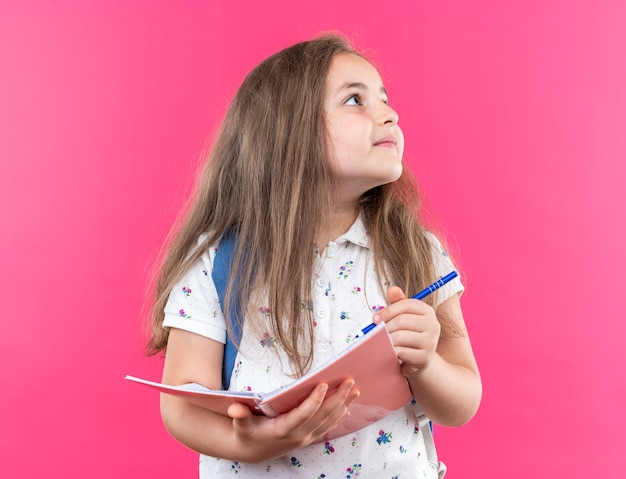 Petite belle fille aux cheveux longs avec sac à dos tenant un cahier et un stylo en levant souriant joyeusement debout sur le rose