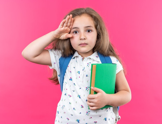 Petite belle fille aux cheveux longs avec sac à dos tenant un cahier avec un visage sérieux saluant debout sur rose