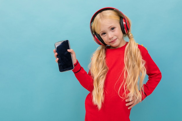 Une petite belle fille aux longs cheveux blonds tressés en queue écouter de la musique avec de gros écouteurs isolé sur fond bleu