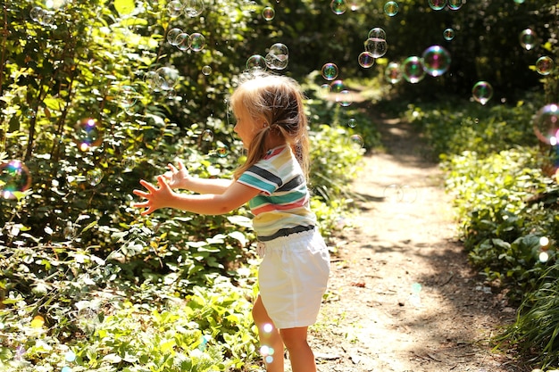 une petite belle fille blonde dans un T-shirt léger et un short blanc attrape des bulles de savon dans le parc