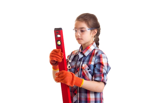 Petite belle fille en chemise à carreaux avec des gants et des lunettes tenant le niveau du bâtiment en studio