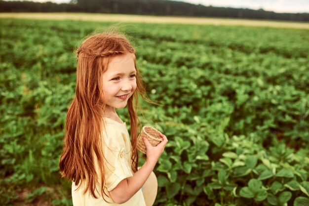 Une petite belle fille dans le champ vert récolte et mange des fraises en s'amusant