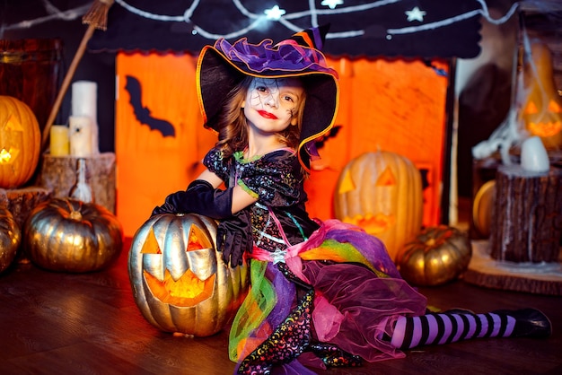 Une petite belle fille dans un costume de sorcière célèbre à la maison dans un intérieur avec des citrouilles et une maison magique en carton sur l'arrière-plan