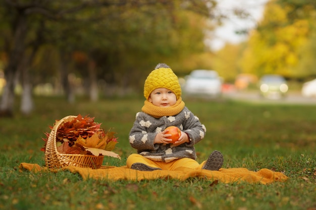 Petite belle fille dans des vêtements chauds et élégants est assis sur un tapis sur la pelouse avec apple assis dans le parc en automne