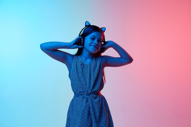 Petite belle fille enfant écoutant de la musique dans des écouteurs sur un studio rose bleu dégradé