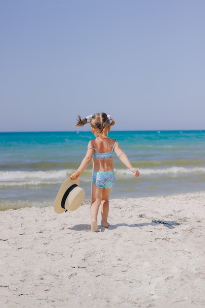 La petite belle fille marche sur la mer sur la plage dans un maillot de bain de sirène et un chapeau de paille