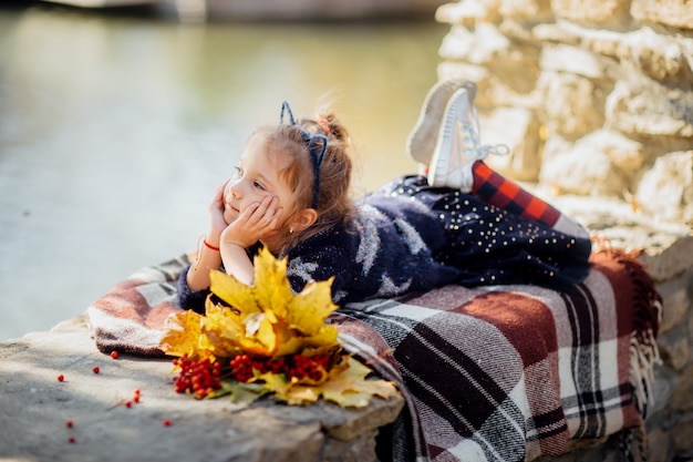 Petite belle fille avec les oreilles du chat se trouve sur le plaid dans le parc en automne avec des feuilles jaunes et Rowan rouge résistant à ses mains et sourires rêveurs
