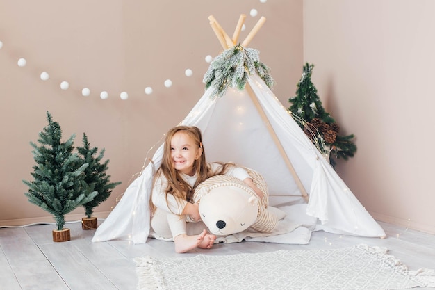 Petite belle fille en pyjama blanc joue avec un ours en peluche près de la chambre des enfants tipi avec c