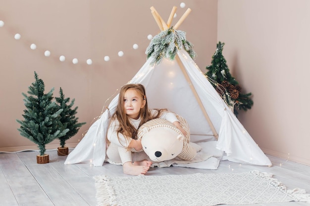 Petite belle fille en pyjama blanc joue avec un ours en peluche près du tipi Chambre des enfants avec décorations et décorations pour arbres de Noël