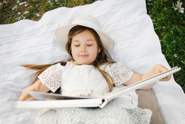 Une petite belle fille se trouve sur une couverture dans le jardin et regarde un album photo de mariage de famille ou de parent