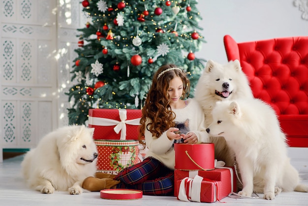 Une petite belle fille et trois grands chiens blancs moelleux à côté de l'arbre de Noël du Nouvel An déballent des boîtes rouges avec des cadeaux. Intérieur festif du nouvel an.