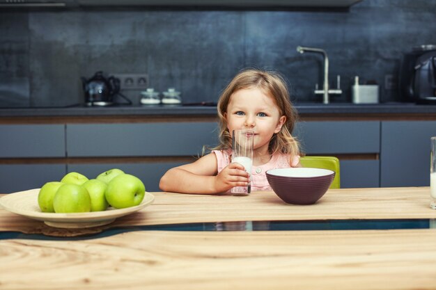 Petite Belle Petite Fille Heureuse à La Maison Dans La Cuisine à La Table Avec Des Pommes Et Du Lait