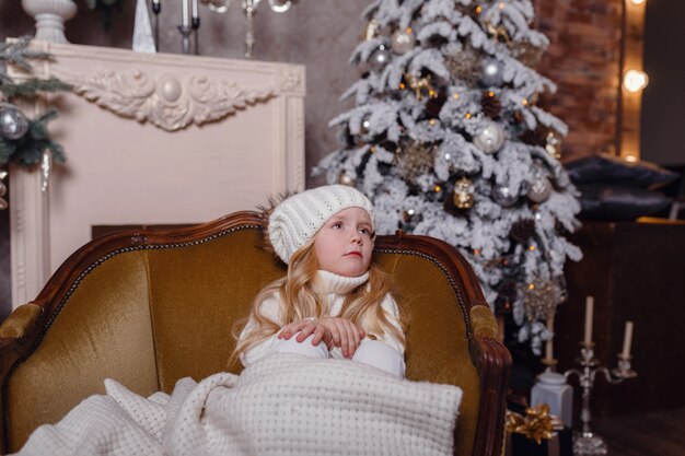 Petite blonde charmante est assise enveloppée dans un tapis dans une belle vieille chaise sous le sapin de Noël.