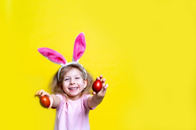 Petite blonde heureuse avec des oreilles de lapin de Pâques sourit et tient des oeufs de Pâques colorés sur fond jaune avec un espace de copie, le concept de la fête religieuse de Pâques.