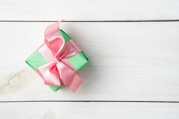 Une petite boîte cadeau dans un emballage vert avec un ruban rose sur un fond en bois blanc.