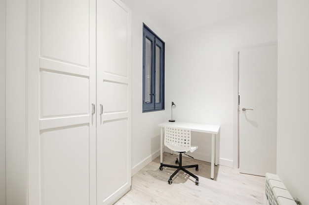 Une petite chambre avec un bureau blanc avec une chaise pivotante assortie et une armoire avec des portes laquées blanches et des poignées en métal