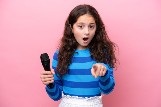 Petite chanteuse prenant un microphone isolé sur fond rose surpris et pointant vers l'avant