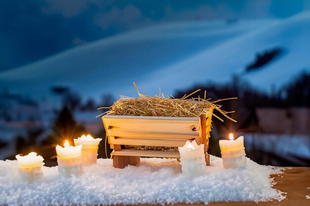Petite crèche en bois remplie de paille sur la neige et des bougies autour et un ciel bleu