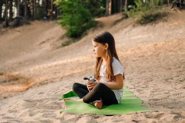 Photo la petite dame s'assoit sur un tapis vert sportif sur le sable et tient une bouteille d'eau fille regarde les haltères de la fiole allongées près de l'enfant