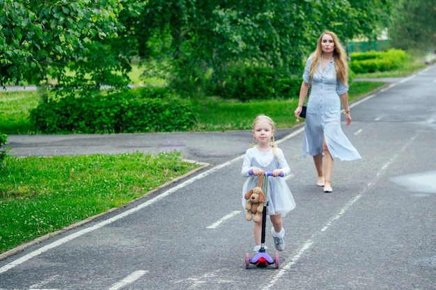 Petite et douce fille blonde dans une jolie robe grise sur un scooter avec une belle mère marchant dans le parc sur fond d'arbres et de verdure