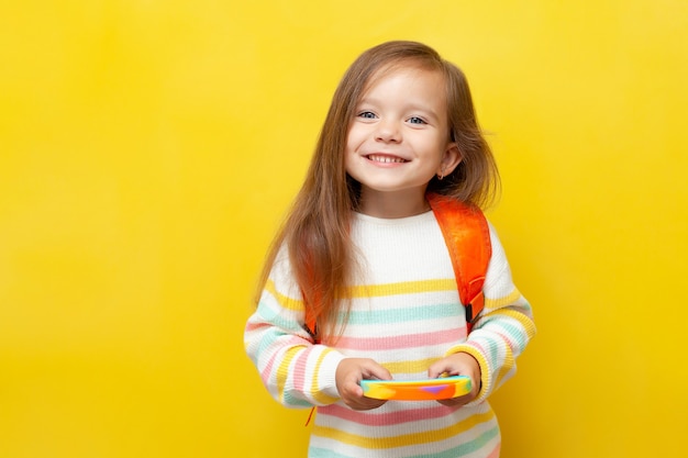 Une petite écolière en blouse rayée et avec un sac à dos tient un pop dans ses mains et sourit