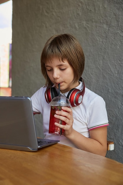 Une petite écolière caucasienne boit de la limonade à travers de la paille utilise un ordinateur portable au café