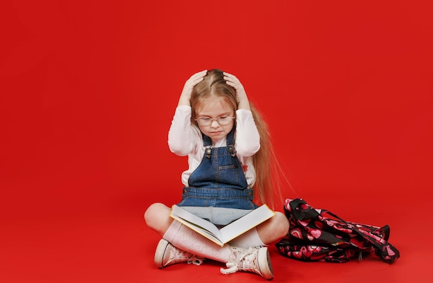 Une petite écolière est assise avec un livre bouleversé avec ses mains sur sa tête, s'inquiétant sur un fond rouge