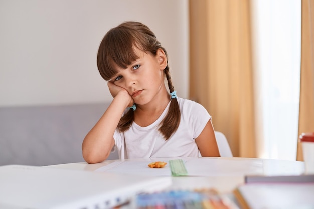 Petite écolière fatiguée et ennuyée, triste, déprimée, souffrant de stress, submergée par la charge de devoirs et de devoirs, assise au bureau, appuyée sur sa main