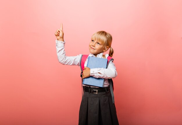 Petite écolière sur fond rose. L'enfant tient un manuel et montre un pouce vers le haut. Notion d'éducation.