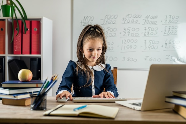 Petite écolière intéressée assise à la maison à la table en regardant une leçon vidéo éducative Curieuse petite fille d'âge scolaire pense à la question de l'enseignant