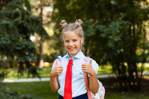 La petite écolière mignonne tient un sac à dos et sourit à la caméra dans le parc.