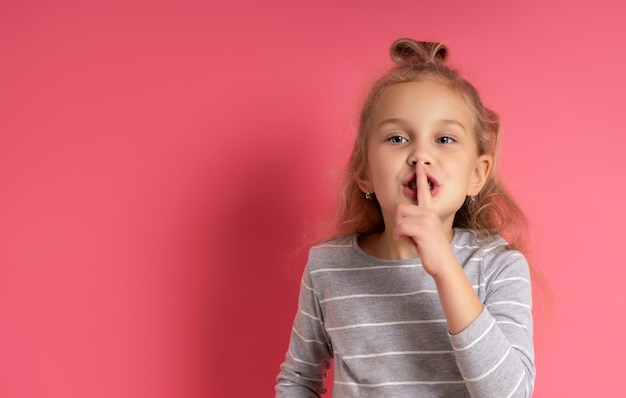 Petite Enfant Blonde Avec Une Coiffure Chignon Vêtue D'un Chemisier à Rayures Grises Elle Montre Un Signe De Silence Posant Sur Fond Rose