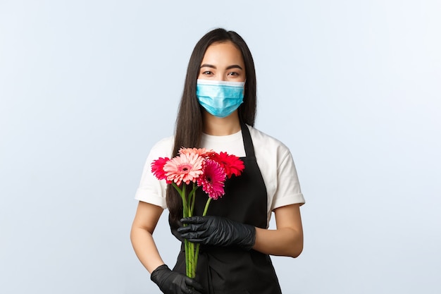 Petite entreprise, pandémie de coronavirus, commandes et concept de magasins. Fleuriste asiatique mignonne souriante en masque médical, tenant un bouquet pour le client, vendant des fleurs pendant l'épidémie de covid-19.