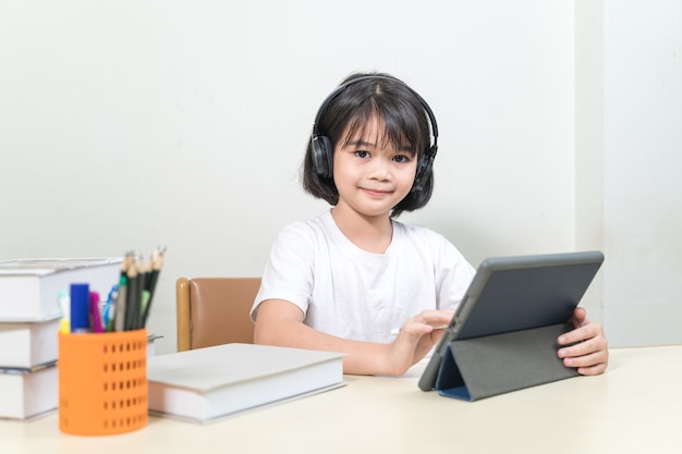 Une petite étudiante asiatique joyeuse porte un casque écrit sur une tablette numérique pour étudier à la maison