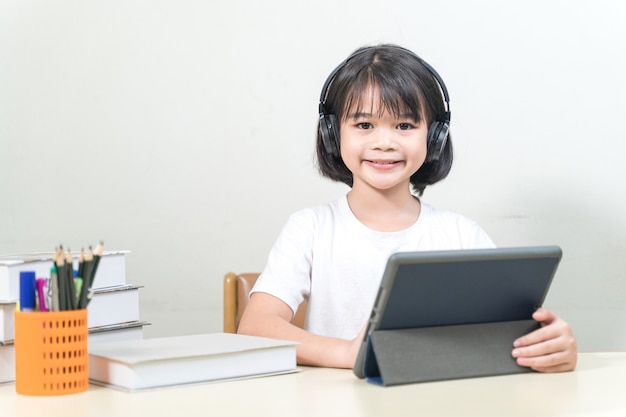 Une petite étudiante asiatique joyeuse porte un casque écrit sur une tablette numérique pour étudier à la maison