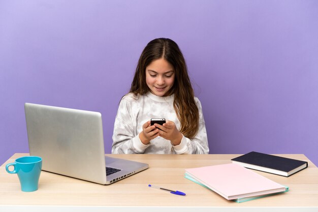 Petite étudiante dans un lieu de travail avec un ordinateur portable isolé sur fond violet envoyant un message avec le mobile