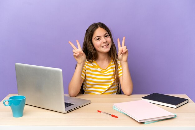 Petite étudiante dans un lieu de travail avec un ordinateur portable isolé sur fond violet montrant le signe de la victoire avec les deux mains