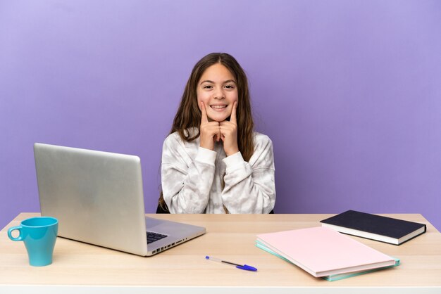 Petite étudiante dans un lieu de travail avec un ordinateur portable isolé sur fond violet souriant avec une expression heureuse et agréable