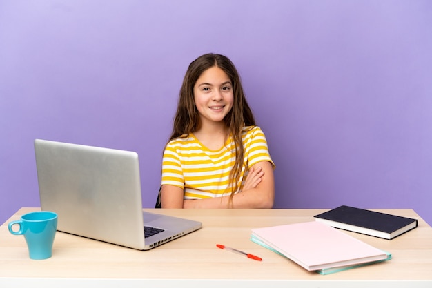 Petite étudiante dans un lieu de travail avec un ordinateur portable isolé sur un mur violet en gardant les bras croisés en position frontale