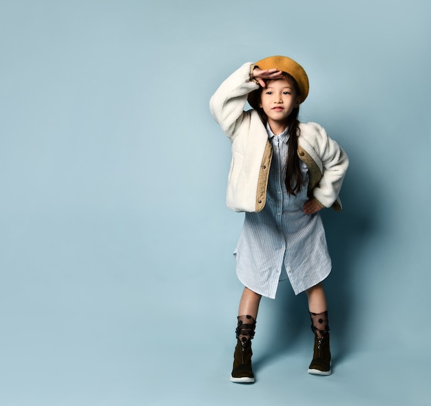 Photo petite femme asiatique en veste, robe chemise, béret marron, bottes. elle sourit, regarde quelqu'un au loin, pose sur fond bleu. enfance, mode, style hipster. pleine longueur, espace de copie