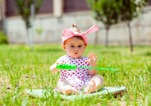 Petite fille de 11 mois est assise dans le parc sur l'herbe. petite fille dans une veste à pois est assise sur l'herbe et joue avec des bulles de savon.