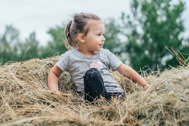 Une petite fille de 3 à 4 ans est assise dans une pile de foin et sourit, sur fond d'arbres