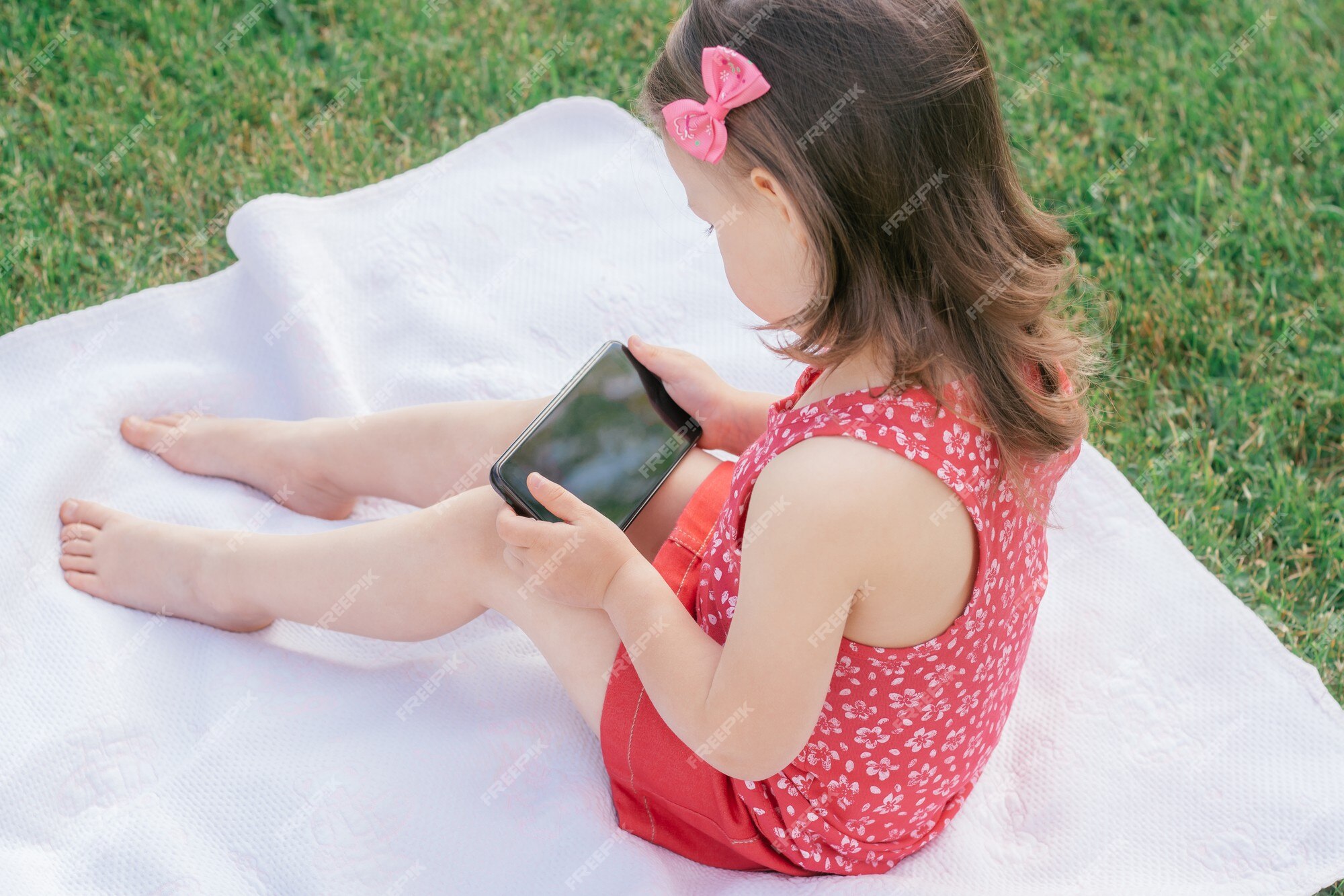 Une Petite Fille De 3 à 4 Ans En Vêtements Rouges Est Assise Sur Une  Couverture Sur L'herbe Verte Et Se Penche Sur Le Téléphone Portable. Enfants,  Utilisant Des Gadgets