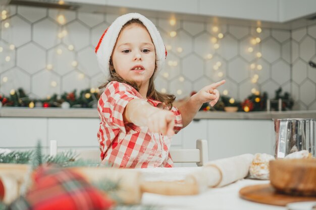 Petite fille de 3 ans en chapeau de Noël rouge et chemise à carreaux prépare la pâte pour les biscuits de pain d'épice dans la cuisine