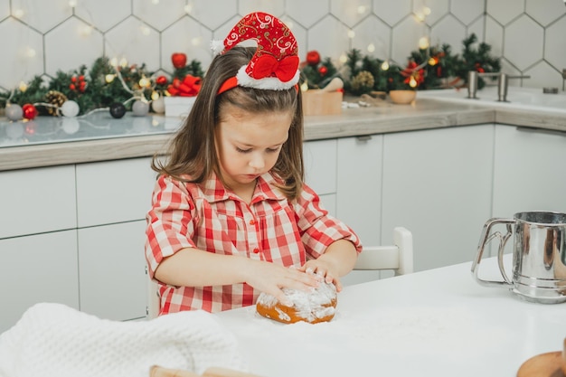 Petite fille de 3 ans en chapeau de Noël rouge et chemise à carreaux prépare la pâte pour les biscuits de pain d'épice dans la cuisine